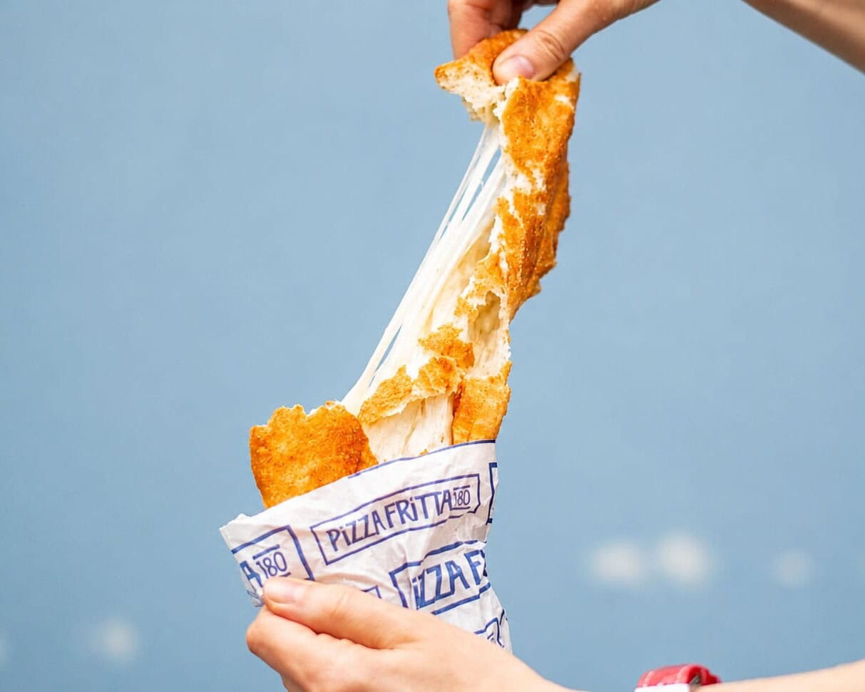 Hands pulling apart a Pizza Fritta 180 fried pizza in its branded wrapper, stretching a dramatic mozzarella cheese pull against a blue sky.