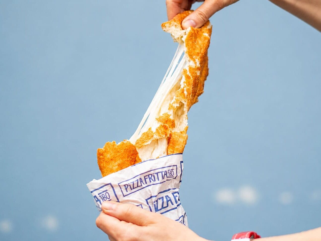 Hands pulling apart a Pizza Fritta 180 fried pizza in its branded wrapper, stretching a dramatic mozzarella cheese pull against a blue sky.