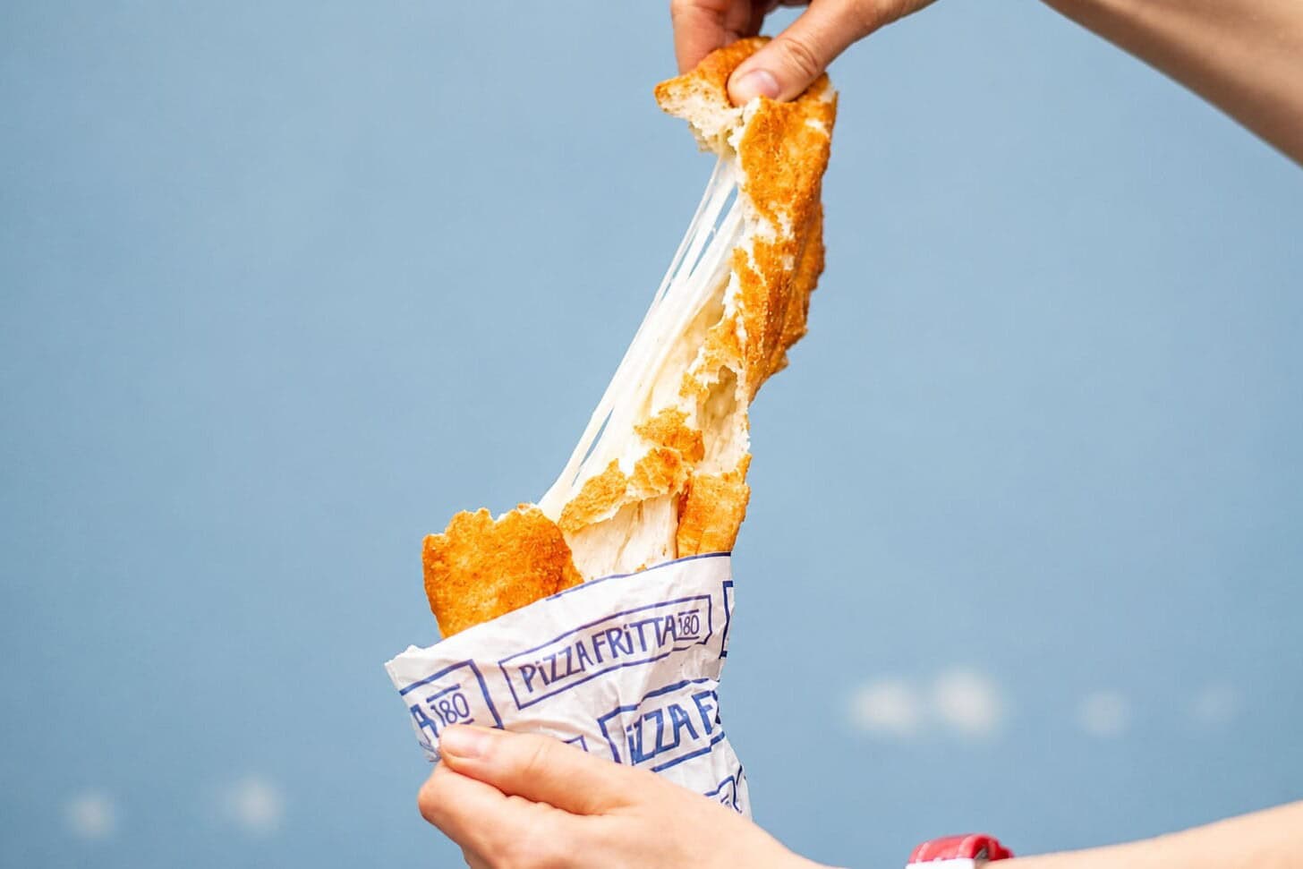 Hands pulling apart a Pizza Fritta 180 fried pizza in its branded wrapper, stretching a dramatic mozzarella cheese pull against a blue sky.