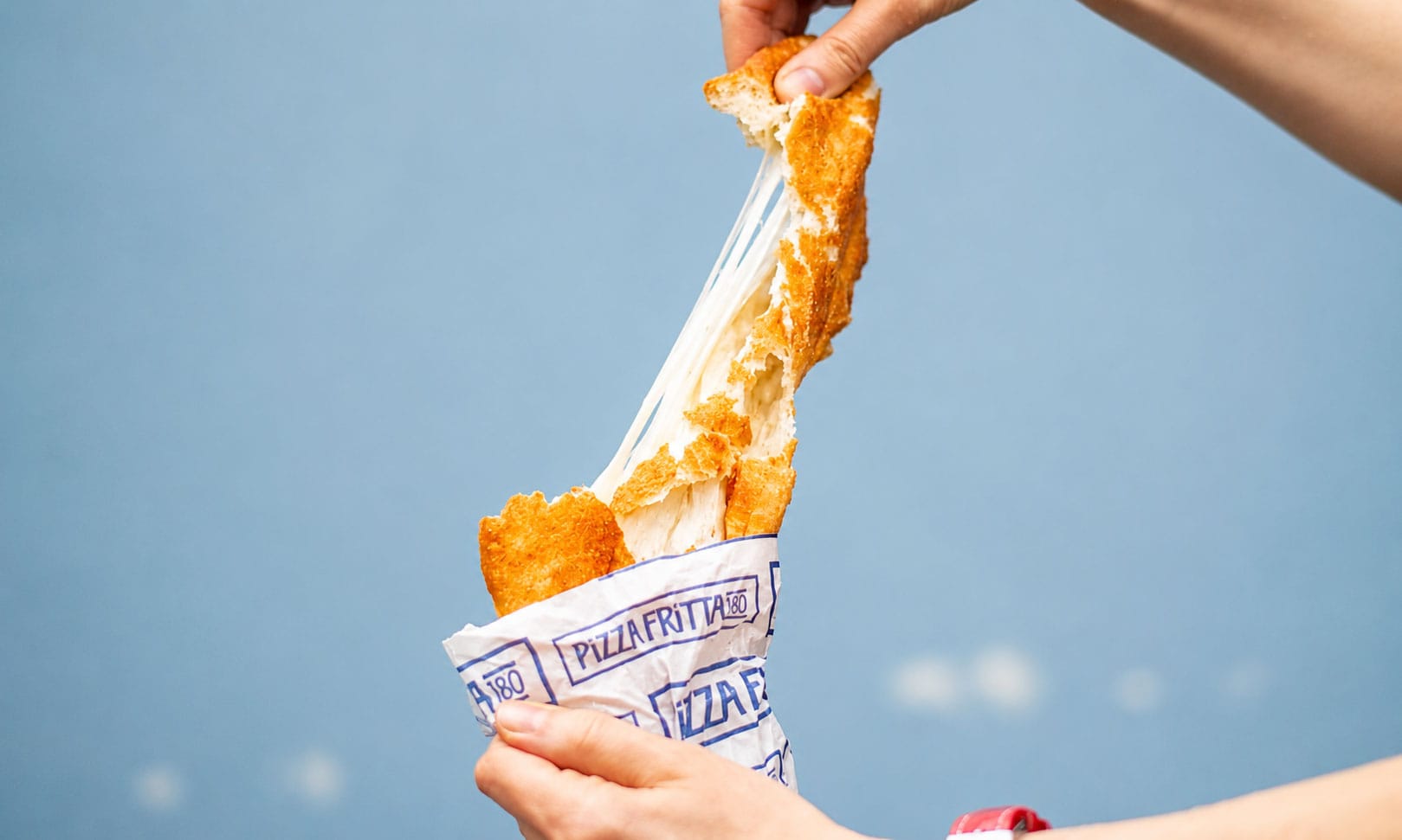 Hands pulling apart a Pizza Fritta 180 fried pizza in its branded wrapper, stretching a dramatic mozzarella cheese pull against a blue sky.