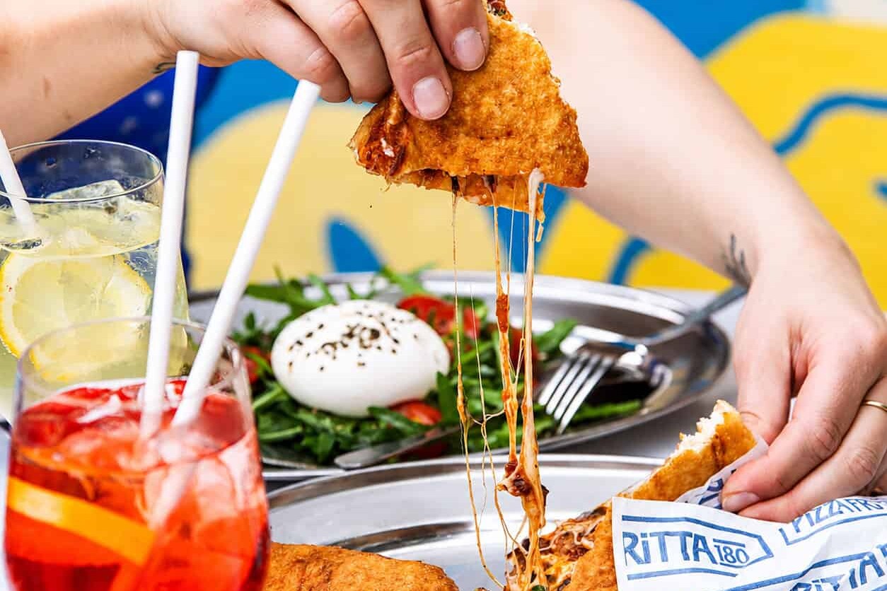 A slice of Pizza Fritta 180 pulled apart to reveal a dramatic cheese pull, served alongside an Aperol Spritz and burrata salad.