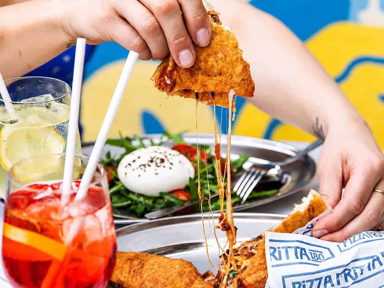 A slice of Pizza Fritta 180 pulled apart to reveal a dramatic cheese pull, served alongside an Aperol Spritz and burrata salad.