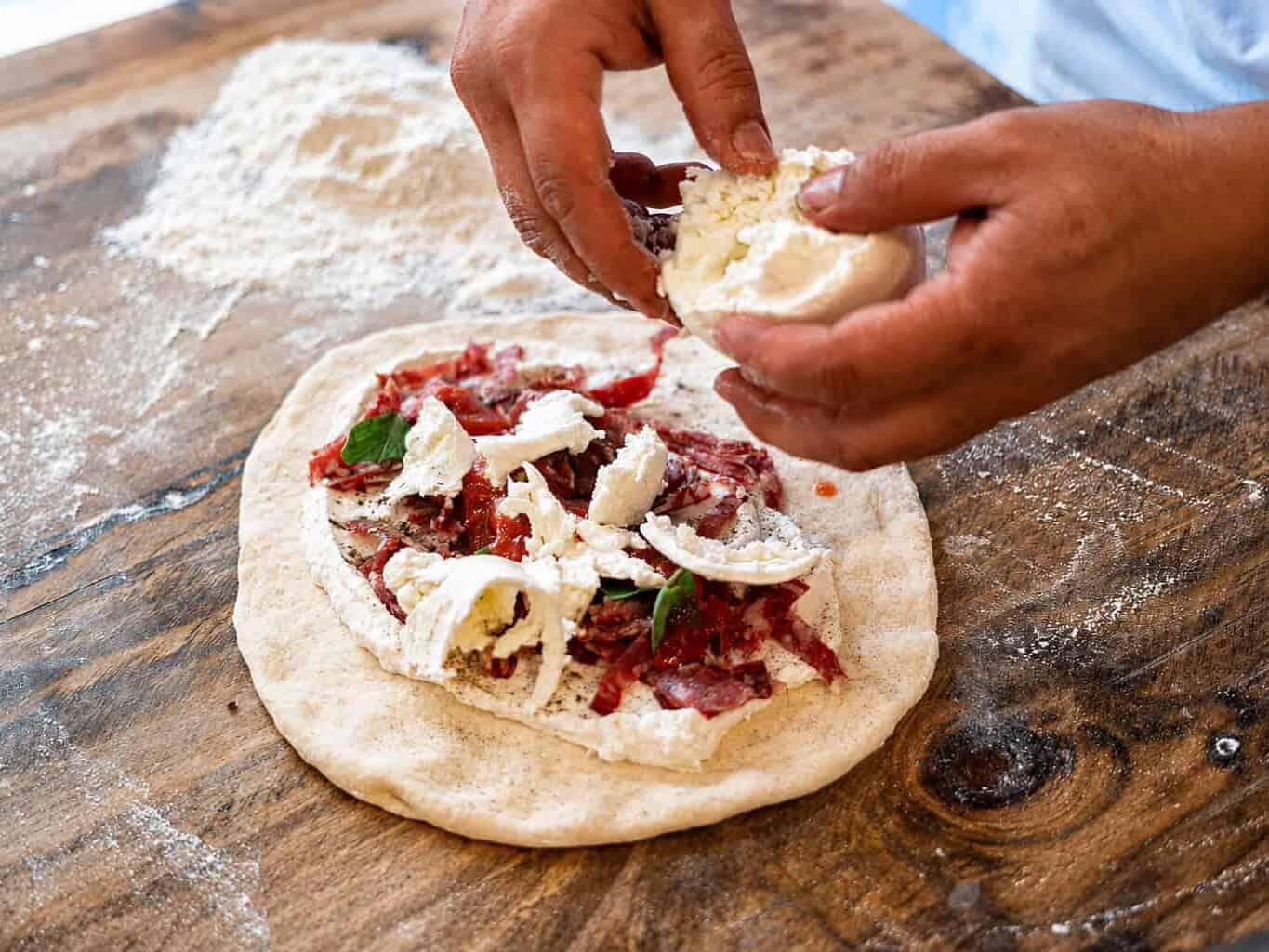 Hands crumbling fresh ricotta onto pizza fritta dough topped with tomato and basil on a floured wooden surface, ready to be folded and fried.