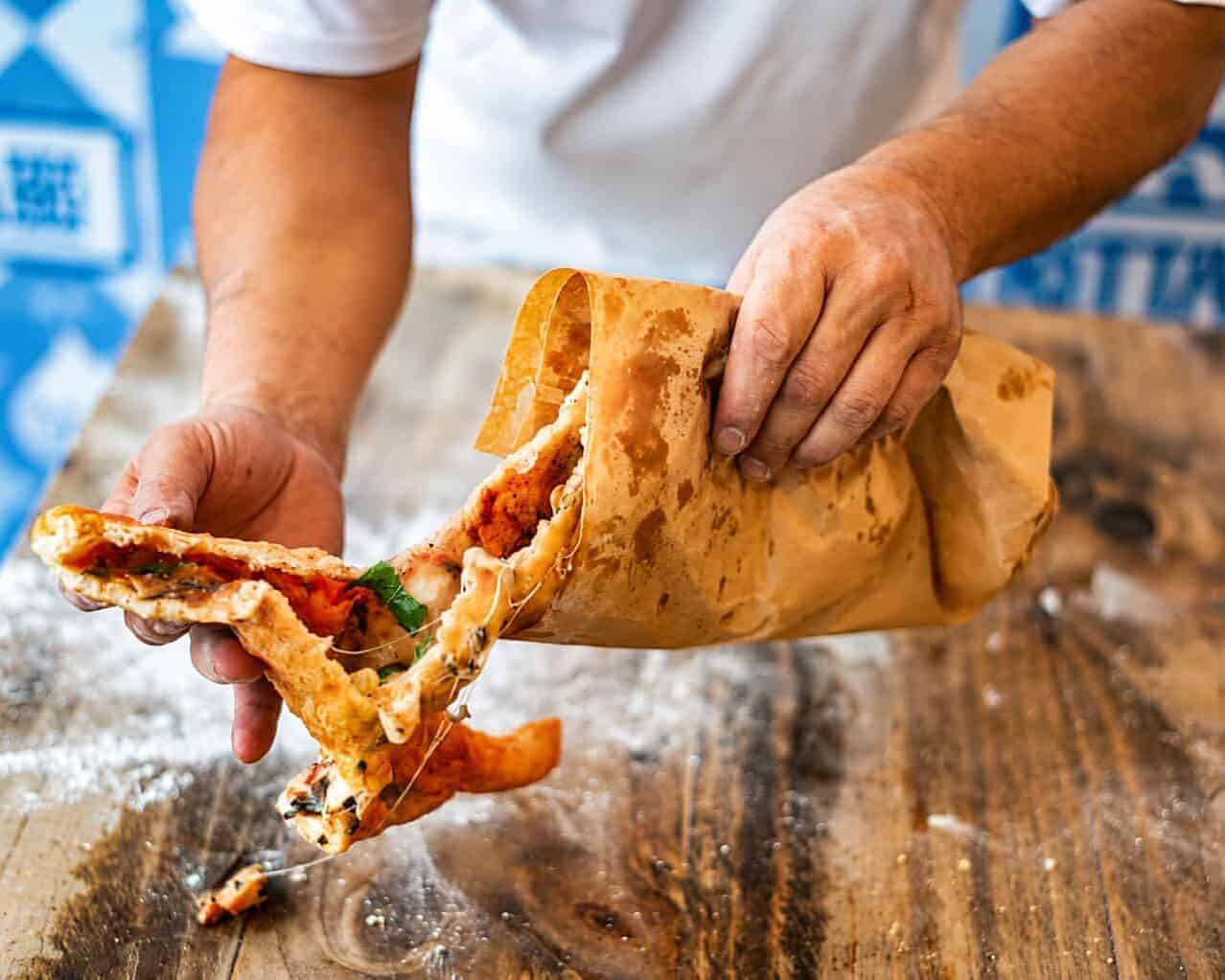 Hands tearing open a crispy pizza fritta to reveal a tomato, mozzarella and basil filling, pulled from its brown paper bag on a floured wooden counter.