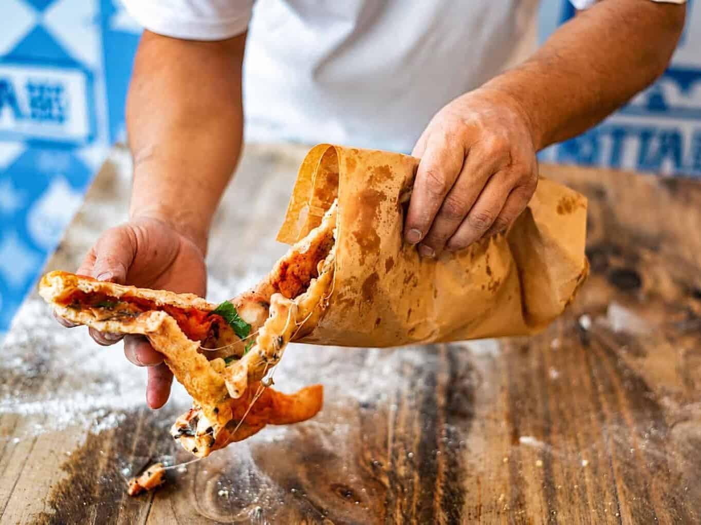Hands tearing open a crispy pizza fritta to reveal a tomato, mozzarella and basil filling, pulled from its brown paper bag on a floured wooden counter.