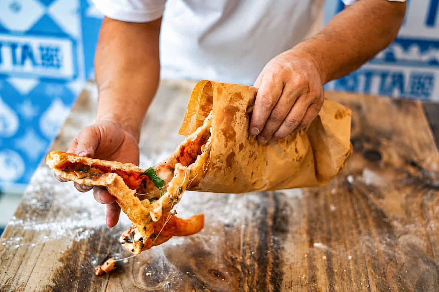 Hands tearing open a crispy pizza fritta to reveal a tomato, mozzarella and basil filling, pulled from its brown paper bag on a floured wooden counter.