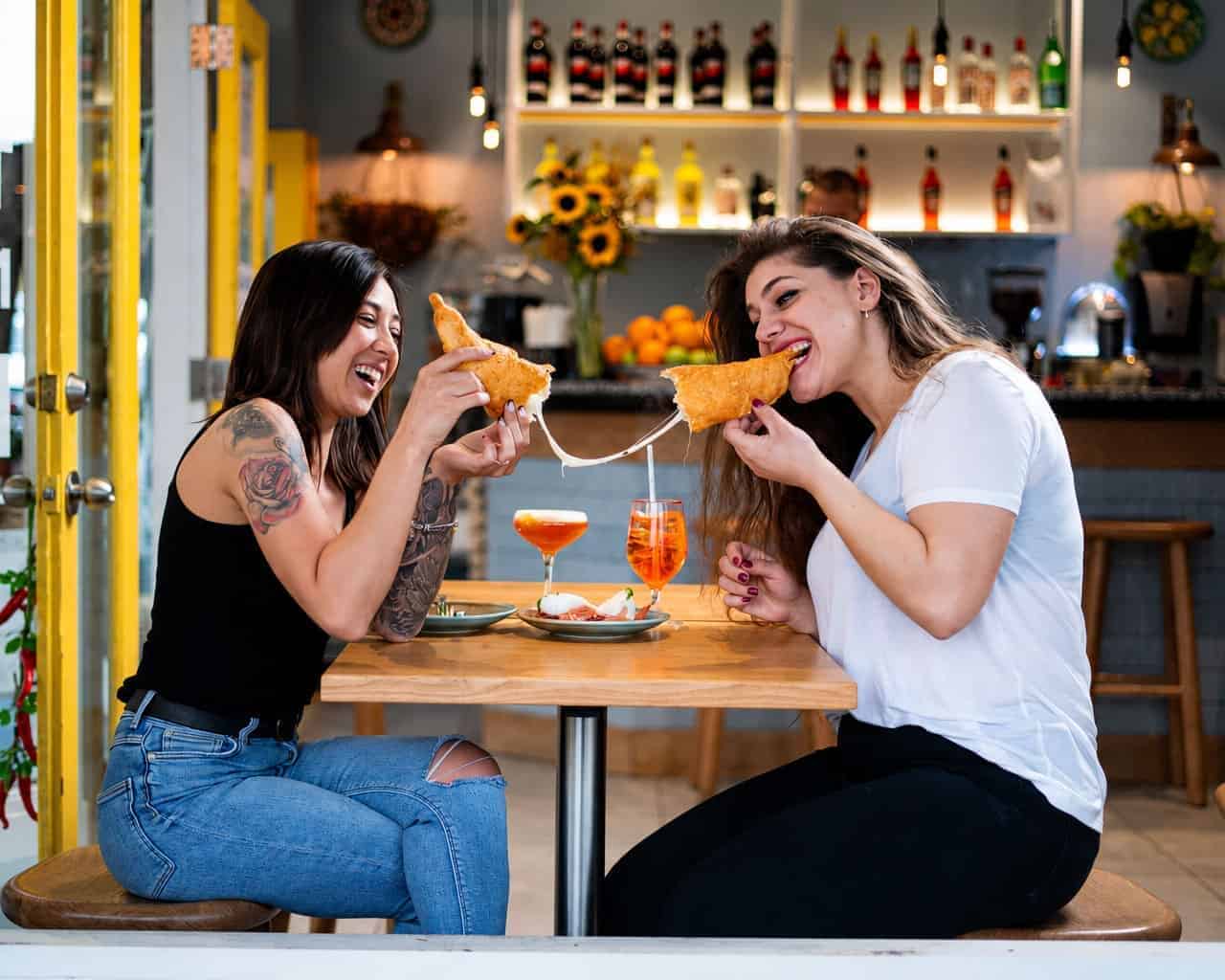 Two women laugh while pulling apart a cheesy pizza fritta at a table inside Pizza Fritta 180, with Aperol Spritzes and a burrata plate in front of them.