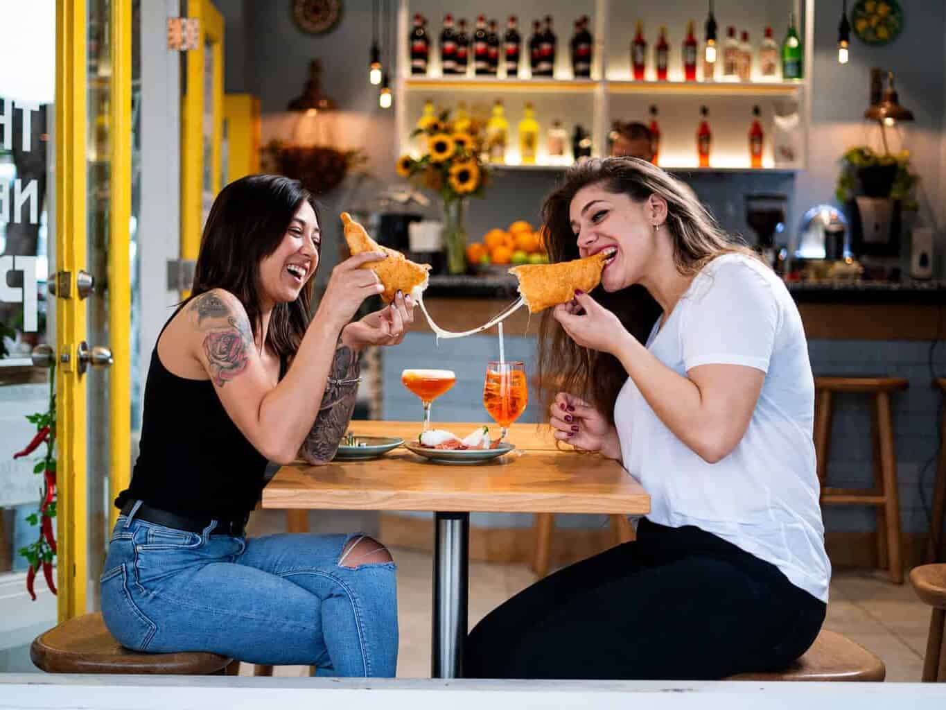 Two women laugh while pulling apart a cheesy pizza fritta at a table inside Pizza Fritta 180, with Aperol Spritzes and a burrata plate in front of them.