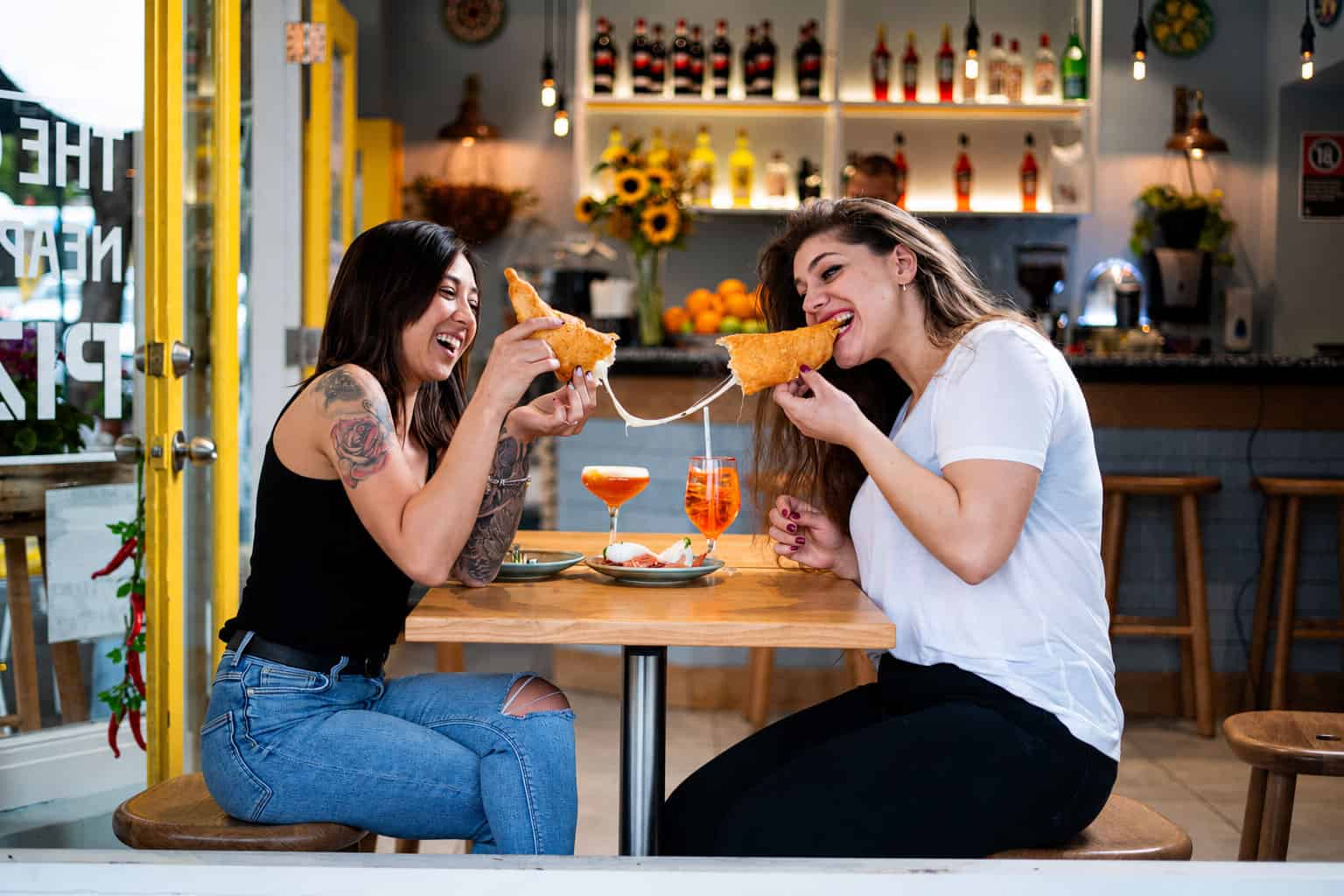 Two women laugh while pulling apart a cheesy pizza fritta at a table inside Pizza Fritta 180, with Aperol Spritzes and a burrata plate in front of them.